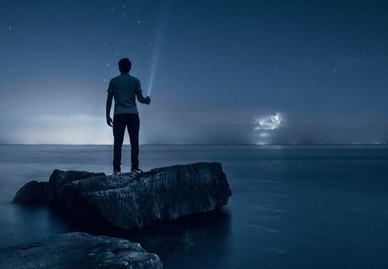Ein Mensch, der im Dunkeln auf einem Felsen steht. Er hat eine Taschenlampe in der Hand, die leuchtet.